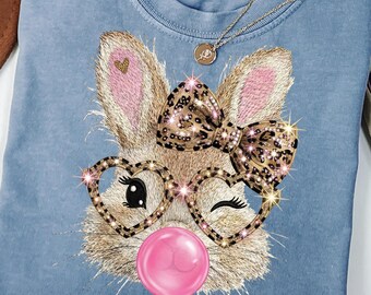 May include: A light blue Comfort Colors t-shirt featuring an embroidered bunny face with heart-shaped glasses, a leopard print bow, and a pink bubble gum bubble. A gold necklace is draped across the shirt.