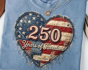 May include: A light blue t-shirt with a heart-shaped design featuring the American flag and the text "250 Years of Freedom 1776-2026." A gold necklace with a pendant rests on the shirt. The shirt is folded.