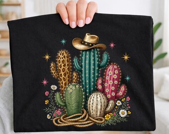 May include: A black t-shirt featuring a graphic print of five cacti in various colors and textures, including leopard print and pink, with a brown cowboy hat resting on the center cactus and a coiled rope at the base, accented by small stars.
