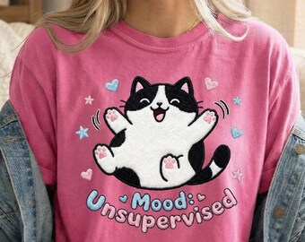May include: A pink t-shirt featuring a cartoon cat with a black and white design, arms outstretched, and the text "Mood: Unsupervised" in blue and pink. The design includes hearts and stars.