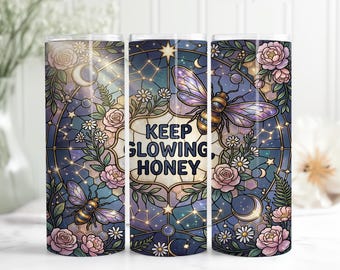 May include: A stainless steel tumbler with a stained glass design featuring bees, flowers, and celestial elements. The tumbler has the text "KEEP GLOWING, HONEY" in a decorative frame. The design includes pink roses, daisies, and a night sky.