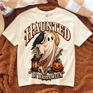 May include: A cream-colored t-shirt featuring a graphic print of a ghost draped in a patchwork quilt, a black raven, a crescent moon, a lit lantern, and a jack-o'-lantern. The text HAUNTED BY HALLOWEEN is printed in a bold, arched font.