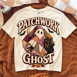 May include: A cream-colored t-shirt featuring a graphic of a ghost draped in a colorful patchwork quilt, accompanied by a black cat with a bow. The text "PATCHWORK GHOST" is printed in large, bold, dark brown letters above and below the illustration.
