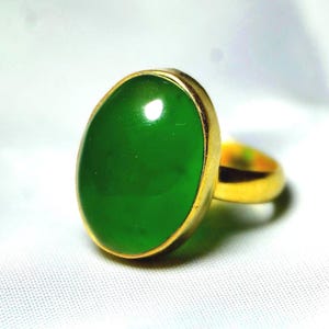 May include: A gold ring featuring a large, oval-shaped, vibrant green gemstone. The gemstone is set in a gold bezel, and the ring band is also gold. The ring is displayed on a white surface.