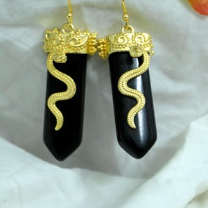 May include: A pair of gold-colored earrings featuring black, elongated pendants. Each earring has a gold-colored snake design wrapping around the black stone. The top of each earring has a decorative gold-colored design. The earrings hang from gold-colored hooks.