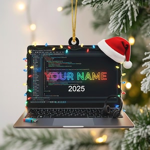 May include: Laptop Christmas ornament with a Santa hat, string lights, and the text "YOUR NAME 2025" in a neon style. The laptop screen displays code. A USB drive and headphones are also visible.