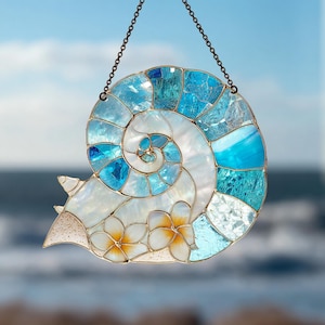 Puede incluir: Atrapasoles de concha marina de vidriera con diseño en espiral en tonos azules y blancos. Cuenta con un contorno de metal dorado y dos flores blancas y amarillas. Incluye una cadena para colgar.