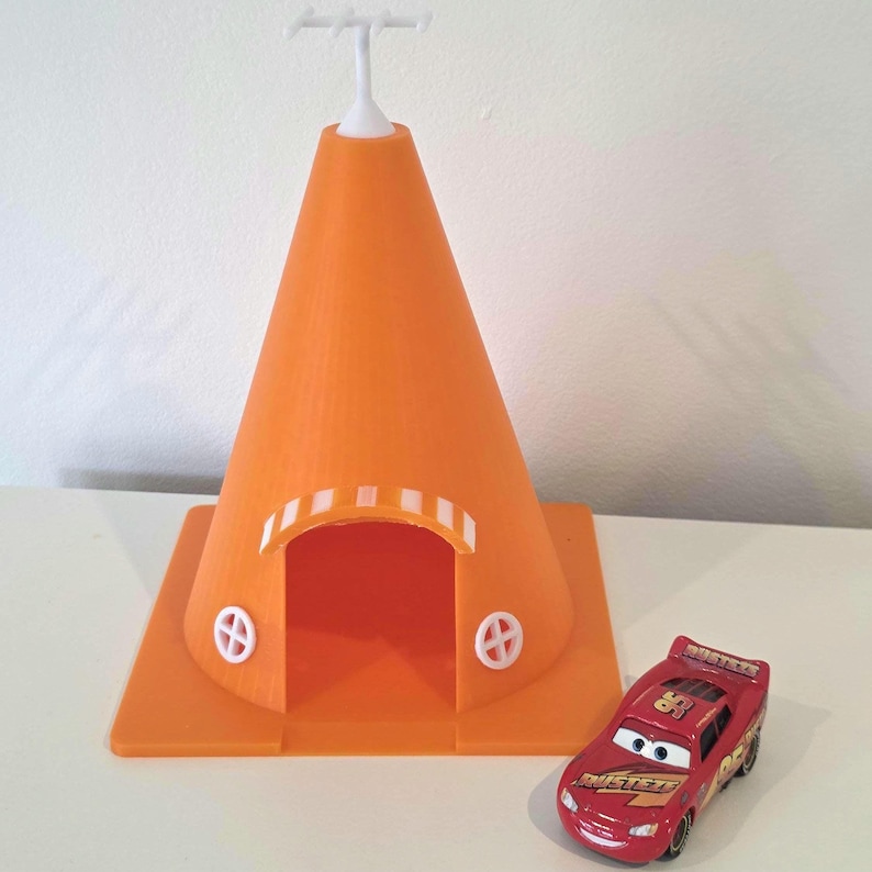May include: An orange 3D-printed cone-shaped structure with a white top and a rectangular base. The structure has a doorway and two circular white accents. A red toy car with the number 95 is next to the structure.