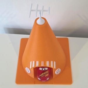 May include: An orange, cone-shaped toy structure with a white antenna on top. A red toy car with the word "Rusteze" is parked inside the structure. The structure has a square base and small, round windows.