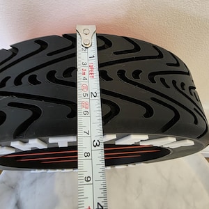 May include: Black tire tread with a patterned design. The tire is being measured with a tape measure, showing a width of approximately 10 cm. The tire has a white and red accent.
