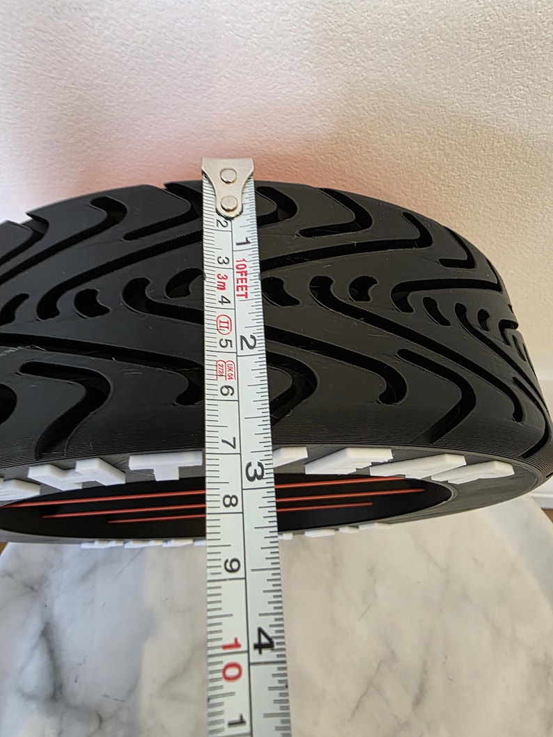 May include: Black tire tread with a patterned design. The tire is being measured with a tape measure, showing a width of approximately 10 cm. The tire has a white and red accent.