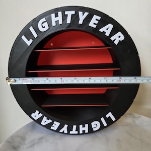 May include: A black and red circular shelf with the word "LIGHTYEAR" in white lettering. The shelf has horizontal red slats and measures approximately 20 cm in diameter.