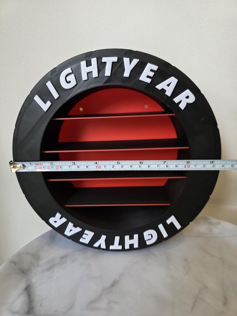 May include: A black and red circular shelf with the word "LIGHTYEAR" in white lettering. The shelf has horizontal red slats and measures approximately 20 cm in diameter.