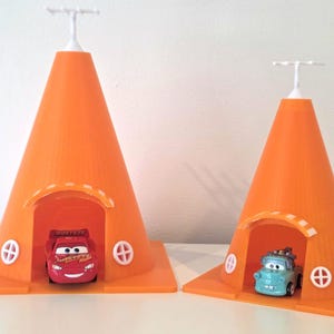 May include: Two orange cone-shaped toy structures with white accents and a white antenna on top. One cone houses a red toy car, the other a blue toy car. The structures have a square base and a small arched opening.