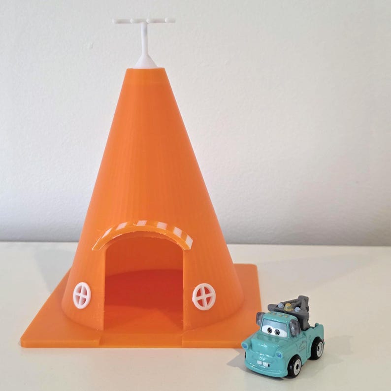 May include: An orange, cone-shaped toy structure with a white top and arched doorway. Two white, circular designs are on the sides. A small, teal-colored toy truck is parked next to the structure.