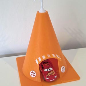May include: An orange 3D-printed toy car shelter with a white antenna and a red toy car inside. The shelter has a cone shape with a rectangular base and small white circular windows.