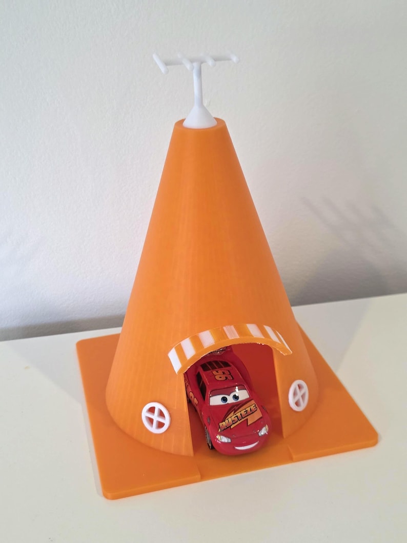 May include: An orange 3D-printed toy car shelter with a white antenna and a red toy car inside. The shelter has a cone shape with a rectangular base and small white circular windows.