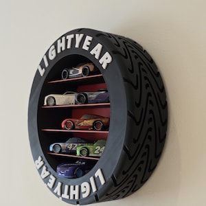 May include: A black, tyre-shaped shelf unit with the word "LIGHTYEAR" in white. It features several levels displaying toy race cars in various colours, including red, blue, and green. The shelf is mounted on a wall.