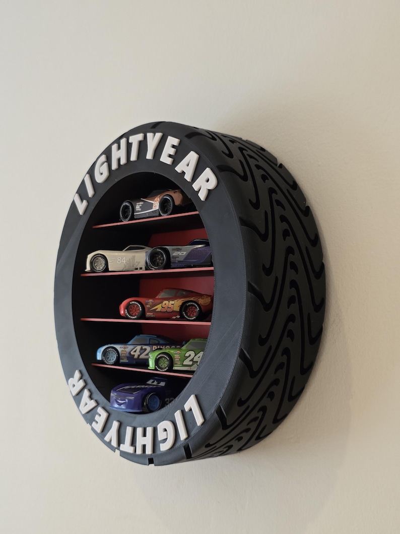 May include: A black, tyre-shaped shelf unit with the word "LIGHTYEAR" in white. It features several levels displaying toy race cars in various colours, including red, blue, and green. The shelf is mounted on a wall.