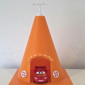May include: An orange 3D-printed toy house with a cone-shaped roof and a square base. The house has two white, circular window designs and a white antenna-like structure on top. A red toy car is inside the house.