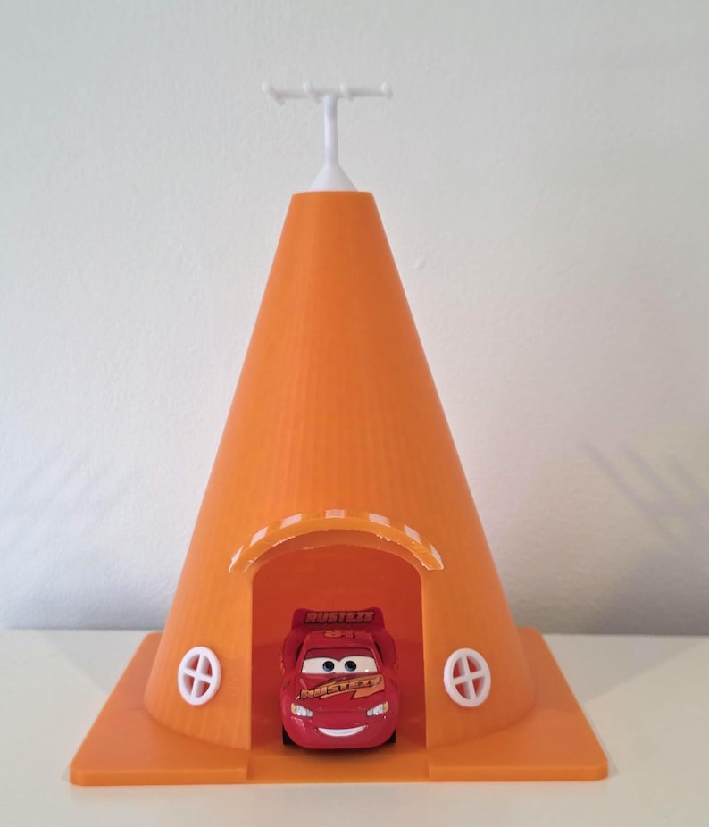 May include: An orange 3D-printed toy house with a cone-shaped roof and a square base. The house has two white, circular window designs and a white antenna-like structure on top. A red toy car is inside the house.