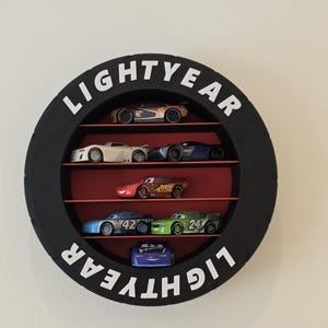 May include: A black, circular shelf with the word "LIGHTYEAR" in white lettering. The shelf contains several toy race cars in various colours, arranged on red shelves. The cars are from the Disney Pixar Cars franchise.
