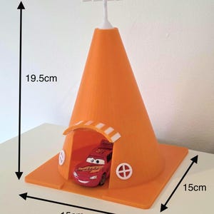 May include: An orange, cone-shaped toy structure with a red race car inside. The structure is 19.5 cm tall and 15 cm wide, with a white top and a small arched opening. The car is red with the number 95.