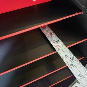 May include: Close-up of a black and red object with a tape measure. The tape measure shows measurements in feet and inches. The object has several horizontal red stripes. The object is approximately 23 cm wide, as measured by the tape.