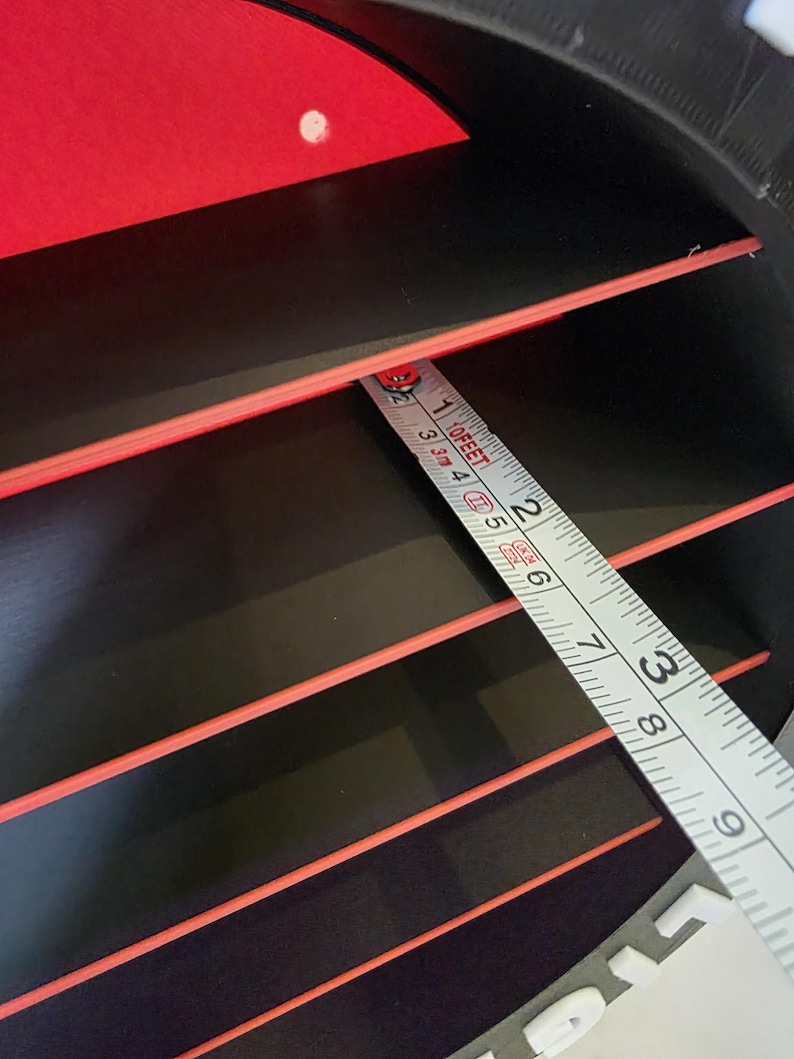 May include: Close-up of a black and red object with a tape measure. The tape measure shows measurements in feet and inches. The object has several horizontal red stripes. The object is approximately 23 cm wide, as measured by the tape.