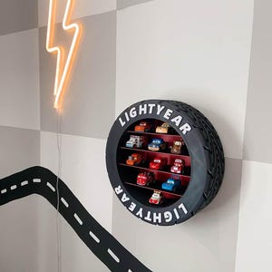 Könnte beinhalten: Ein kreisförmiges Wandregal in Form eines schwarzen Reifens mit rotem Innenraum, das oben und unten den weißen Schriftzug "LIGHTYEAR" trägt. Das Regal enthält zehn kleine Spielzeugautos, die auf drei horizontalen Ebenen angeordnet sind.
