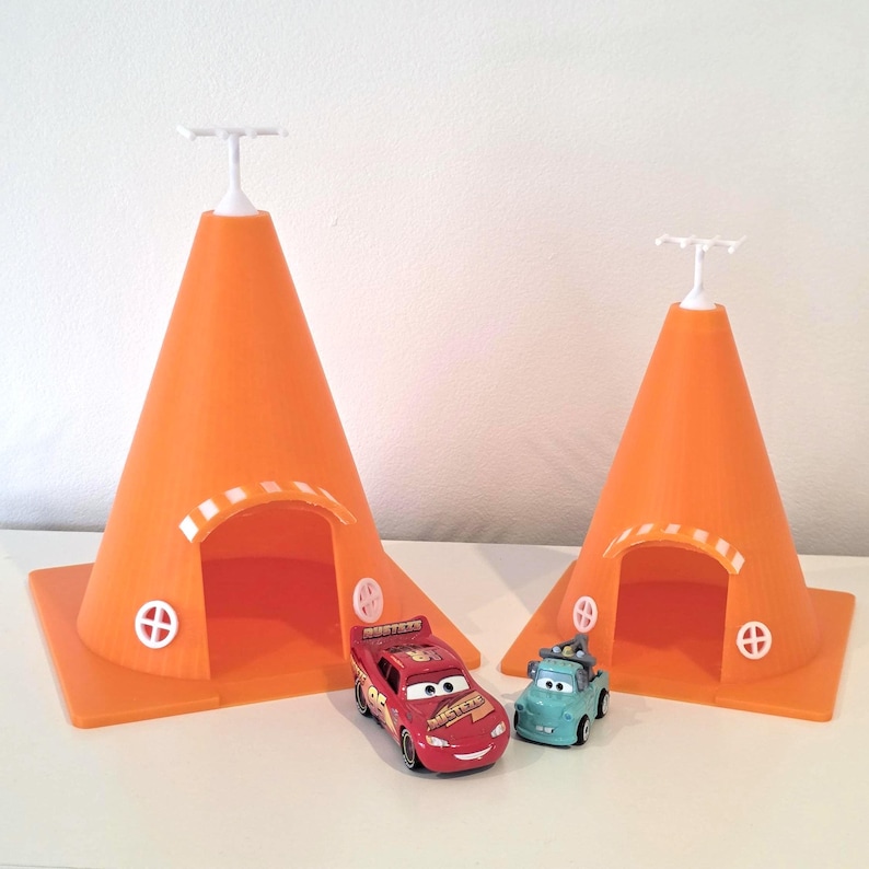 May include: Two orange cone-shaped structures with white accents and small doors, resembling buildings. A red race car and a teal tow truck toy are positioned in front of the structures. The toys are from the Cars movie franchise.