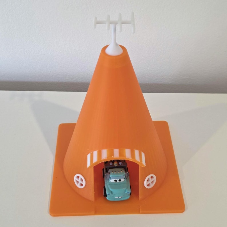 May include: An orange 3D-printed toy based on the Cars movie. The toy features a large traffic cone shape with a small teal car inside. The cone has a white antenna on top and a square base with decorative elements.