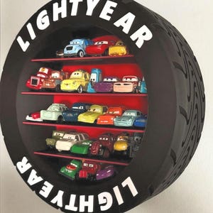 May include: A black, tyre-shaped shelf unit with the word "LIGHTYEAR" in white. It features three red shelves displaying toy cars in various colours. The tyre has a tread pattern.