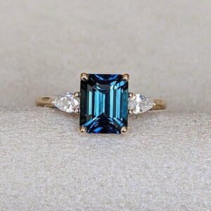 May include: A gold ring featuring a large, rectangular emerald-cut dark blue gemstone at the center, flanked by two smaller, clear pear-shaped stones on each side. The setting is a classic prong style, highlighting the deep blue color of the center stone.