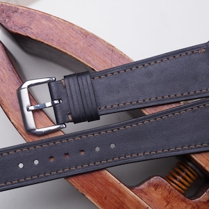 May include: A black leather watch strap with brown stitching and a silver buckle. The strap is displayed on a wooden stand. The watch band has multiple holes for adjustment.