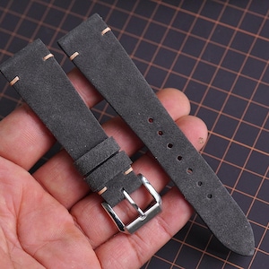 Könnte beinhalten: Zweiteiliges Uhrenarmband aus dunkelgrauem, Wildleder-ähnlichem Material. Das Armband hat hellbeige Nähte und eine silberfarbene Schnalle. Das Uhrenarmband wird vor einem dunklen Hintergrund gezeigt.