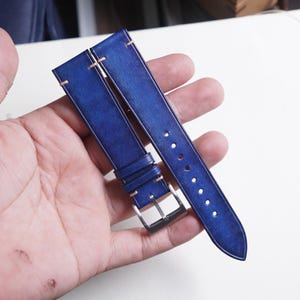 May include: A pair of blue leather watch straps with contrasting stitching. The straps have a silver buckle and are held in a hand. The leather has a distressed appearance.