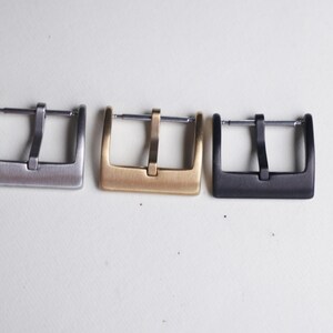 May include: Three watch buckles in a row, each with a rectangular frame and a central bar. The buckles are in silver, gold, and black finishes. The watch buckles are displayed on a white surface.
