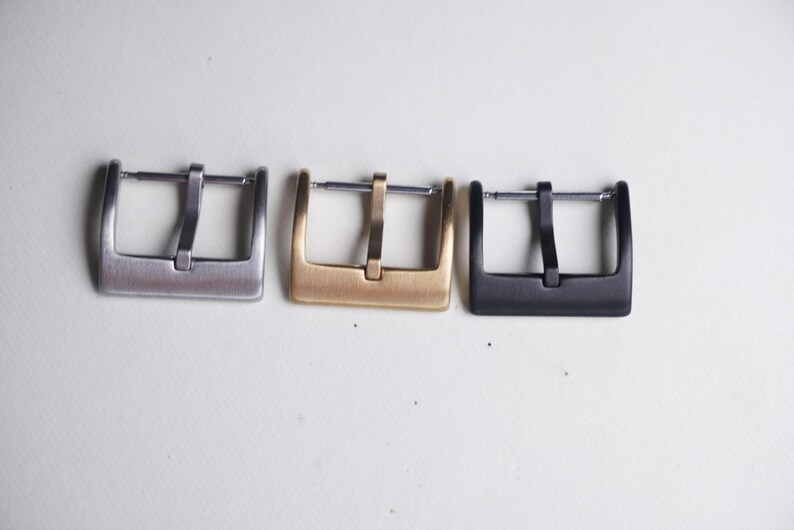 May include: Three watch buckles in a row, each with a rectangular frame and a central bar. The buckles are in silver, gold, and black finishes. The watch buckles are displayed on a white surface.