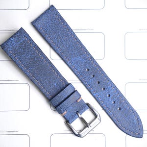 May include: Two blue leather watch straps with a textured finish and light brown stitching. One strap has a silver buckle and a light pink accent. The straps are displayed on a white background with a grid pattern.