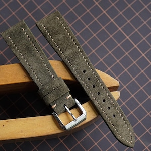 Könnte beinhalten: Olivgrüne Wildleder-Uhrenarmbänder mit weißen Nähten und einer silbernen Schnalle. Die Armbänder werden auf einem hölzernen Uhrmacherwerkzeug präsentiert, das die Textur und die Details des Leders hervorhebt. Der Hintergrund ist ein dunkles Gitter.