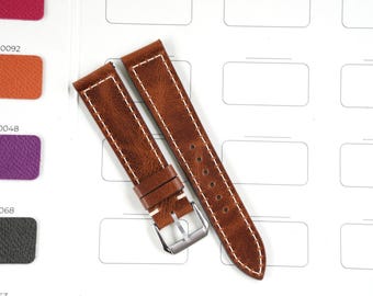 Puede incluir: Dos correas de reloj de cuero marrón con costuras blancas. Las correas tienen una textura envejecida y una hebilla plateada. Las correas están sobre una superficie blanca con muestras de color en el fondo.