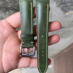 Puede incluir: Correa de reloj de cuero verde oliva con costuras blancas. Cuenta con una hebilla plateada y varios orificios de ajuste. El cuero tiene una apariencia texturizada.