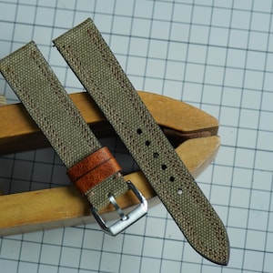 May include: Two olive green canvas watch straps with brown leather accents and silver buckles. The straps are displayed on a wooden tool, showcasing their texture and design. The straps are approximately 0.75 inches wide.