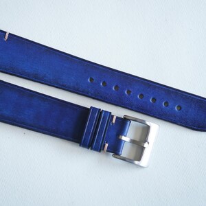 May include: Two-piece watch strap in a deep blue hue. The leather strap features light pink stitching and a silver-colored buckle. The strap has several holes for adjustment and a pointed end.