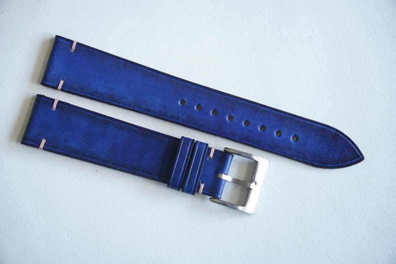 May include: Two-piece watch strap in a deep blue hue. The leather strap features light pink stitching and a silver-colored buckle. The strap has several holes for adjustment and a pointed end.