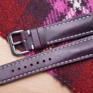 Può includere: Un cinturino per orologio in pelle color prugna con cuciture bianche e fibbia argentata. Il cinturino ha diversi fori di regolazione. Il cinturino dell'orologio è esposto su un tessuto fantasia.
