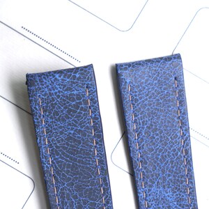 May include: Two blue leather watch straps with a textured, cracked appearance. Each strap has a light brown stitch running down the center. The straps are set against a white background with faint gray lines.