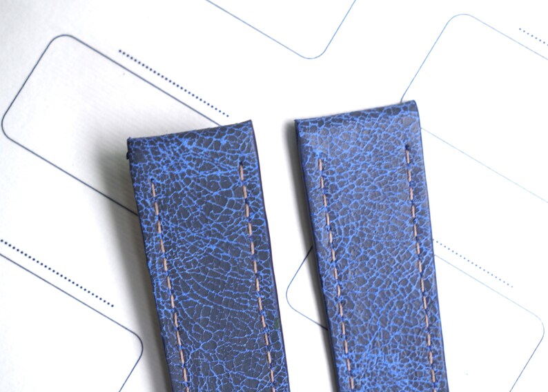 May include: Two blue leather watch straps with a textured, cracked appearance. Each strap has a light brown stitch running down the center. The straps are set against a white background with faint gray lines.