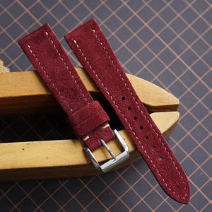 Könnte beinhalten: Burgunderfarbenes Wildleder-Uhrenarmband mit weißen Nähten und einer silbernen Schnalle. Das Armband wird auf einem hölzernen Uhrmacherwerkzeug präsentiert, das seine Textur und sein Design hervorhebt. Das Armband ist ca. 20 cm lang.